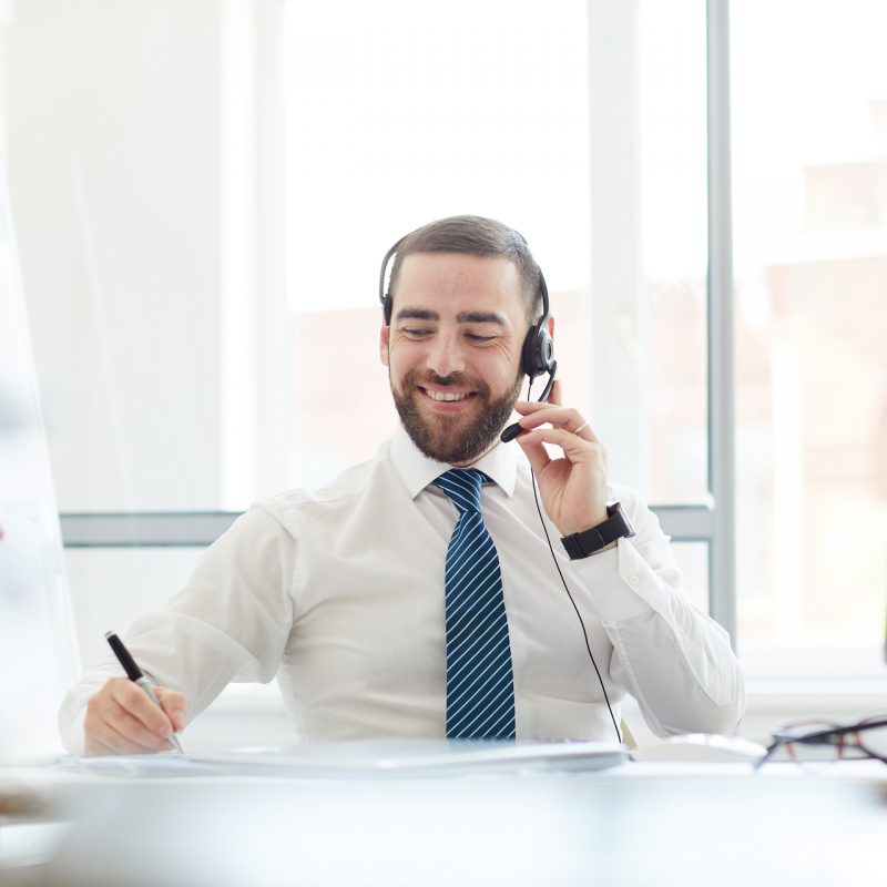 Positive communicative handsome young call center agent sitting at table and making notes in notepad while talking to customer via headset